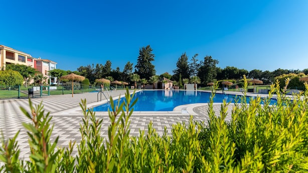 Une piscine entourée d’une terrasse dallée, avec un bâtiment en arrière-plan et une végétation luxuriante.