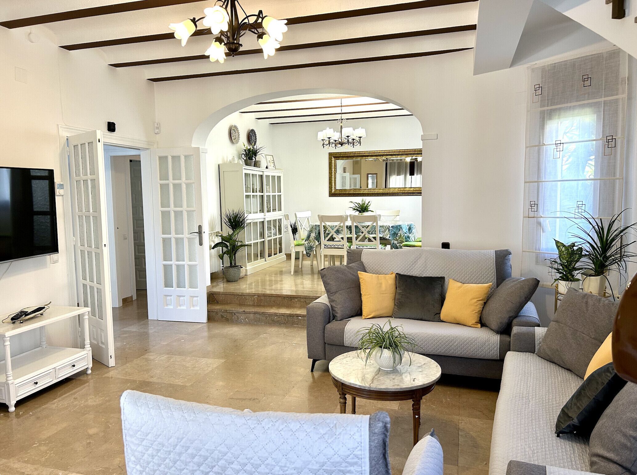 A spacious living room with a grey sofa, a white armchair, and a marble coffee table. There is a TV on the left and a dining area in the background.