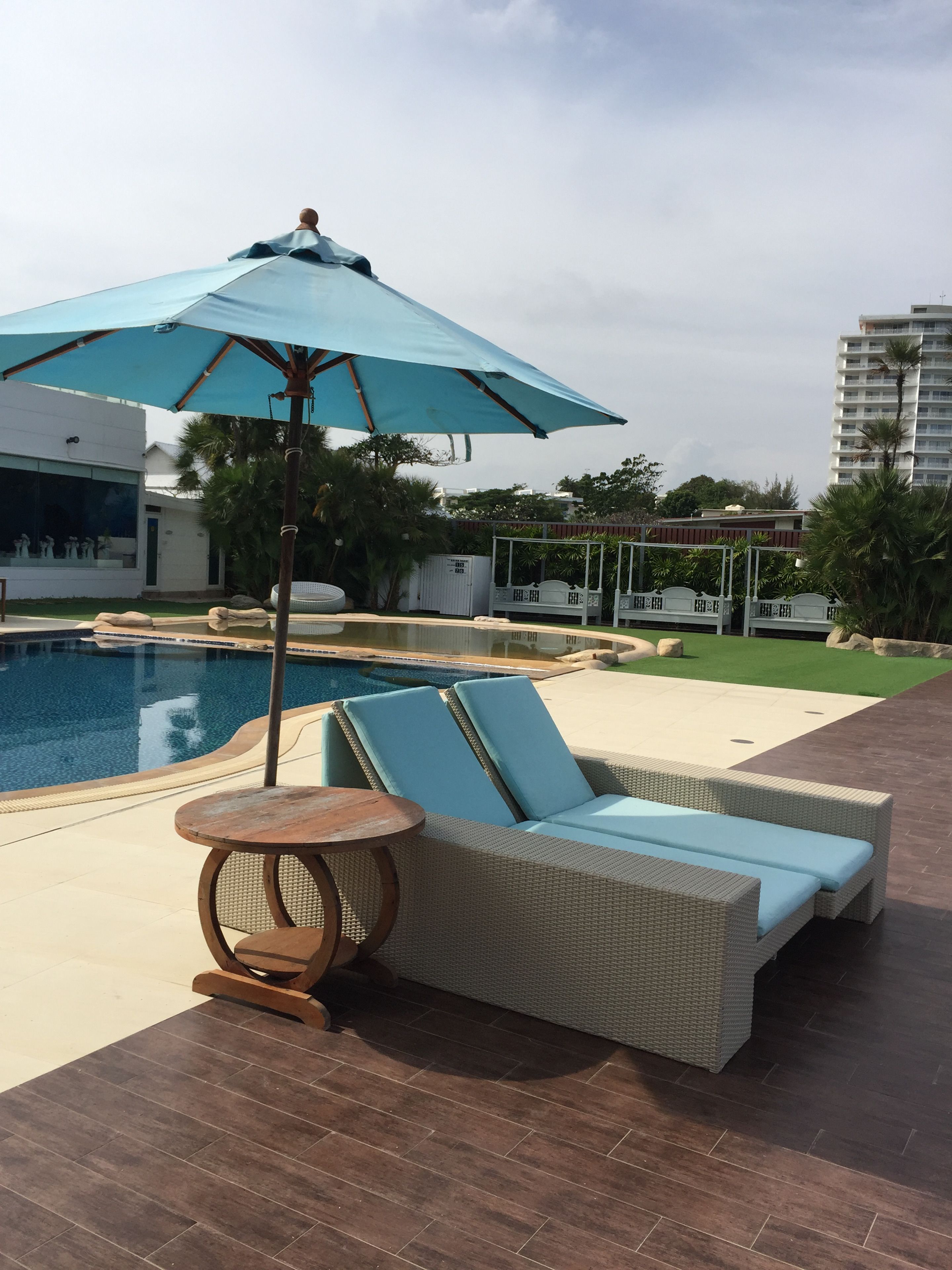 A poolside area with a lounge chair and umbrella, a small table, and a view of a building in the background.