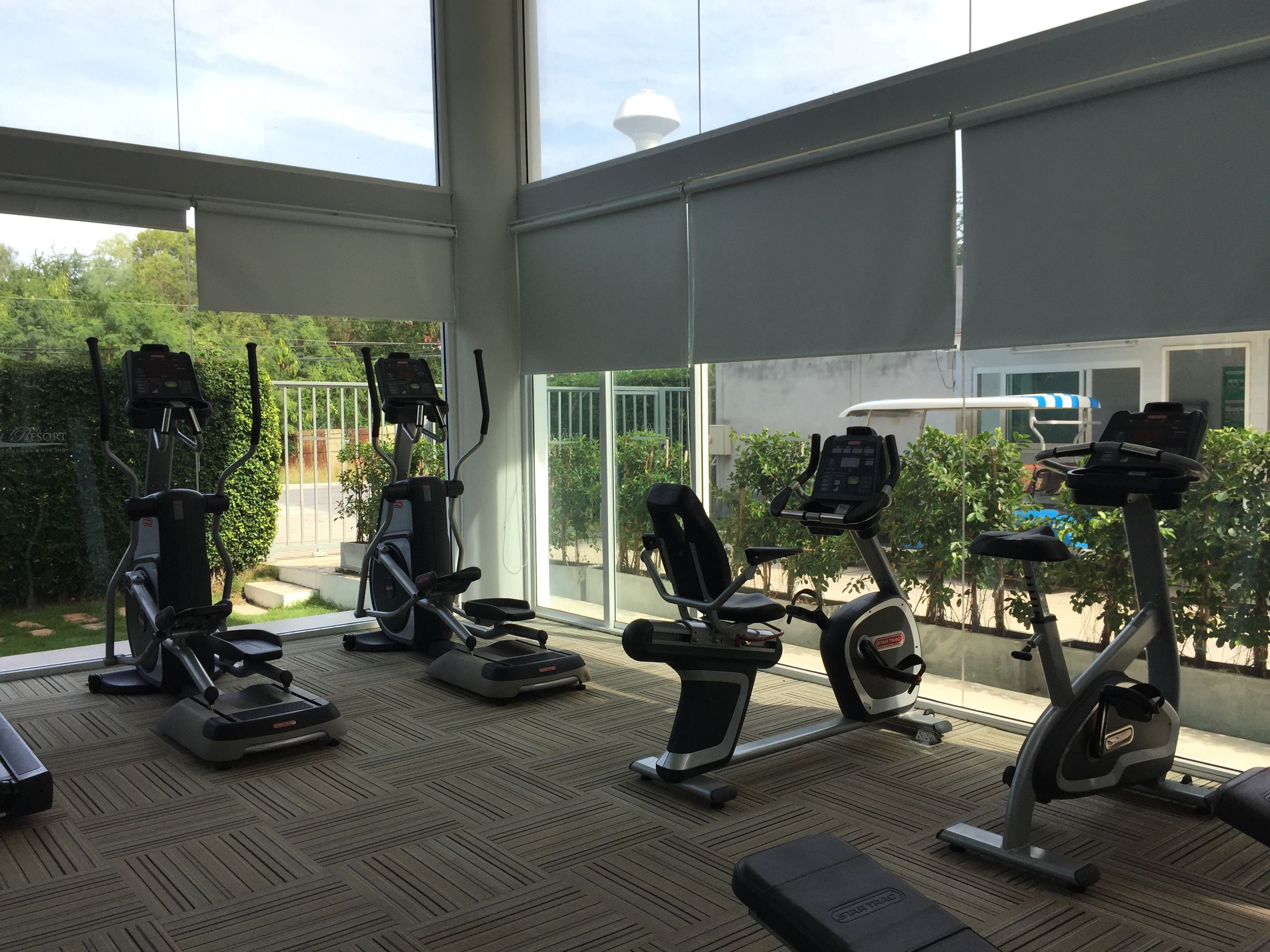 A gym with various exercise machines, large windows, and a view of greenery outside.