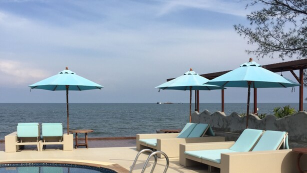 Uma área à beira da piscina com espreguiçadeiras e guarda-sóis, piscina e vista para o mar.
