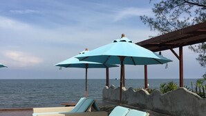 Uma área de estar à beira-mar com espreguiçadeiras de vime e uma pequena mesa, protegida por grandes guarda-sóis, com vista para o mar.