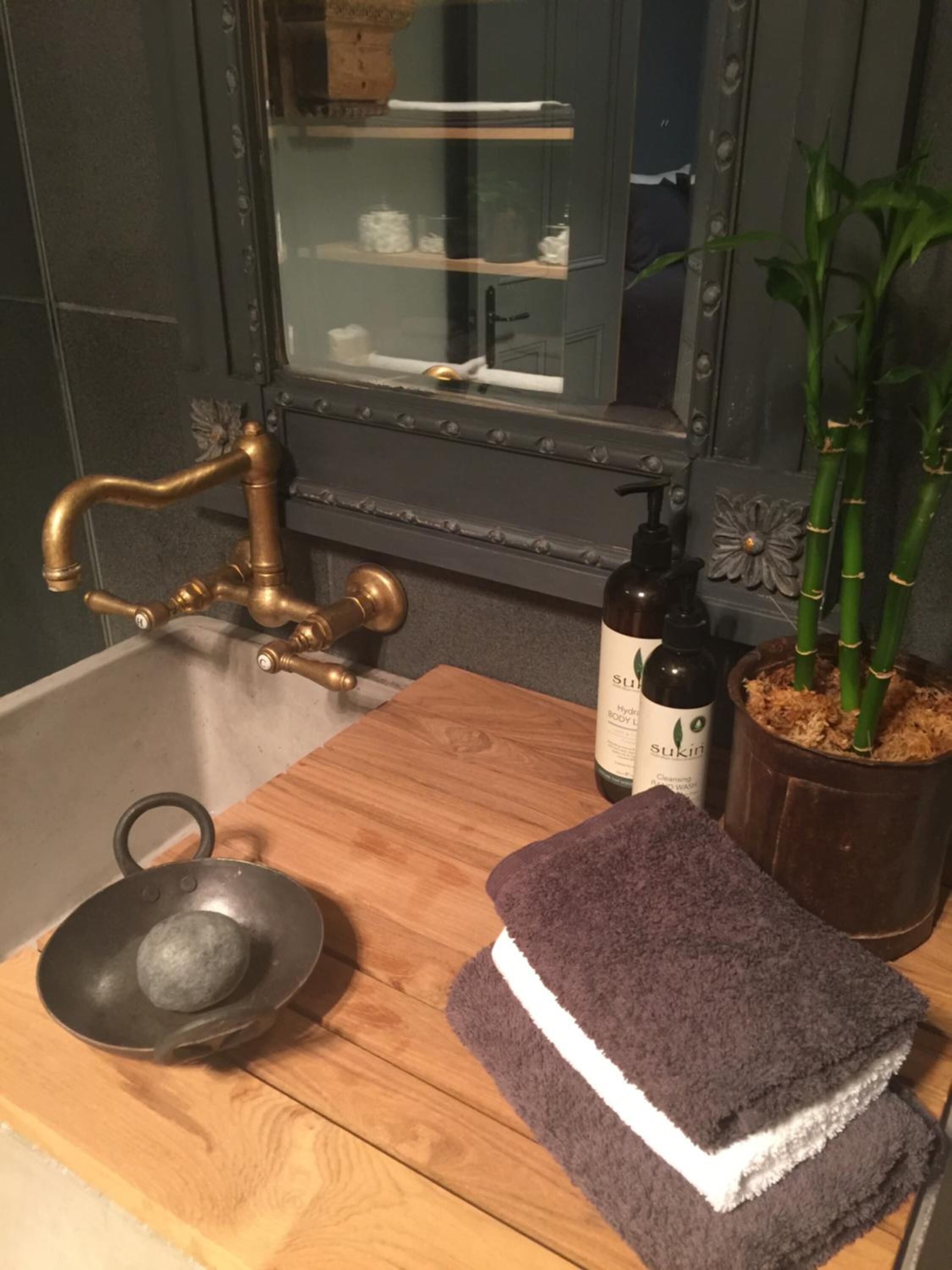 Un baño con tocador de madera, un lavabo, un espejo, una planta de bambú y una botella de Sukin para el cuerpo.