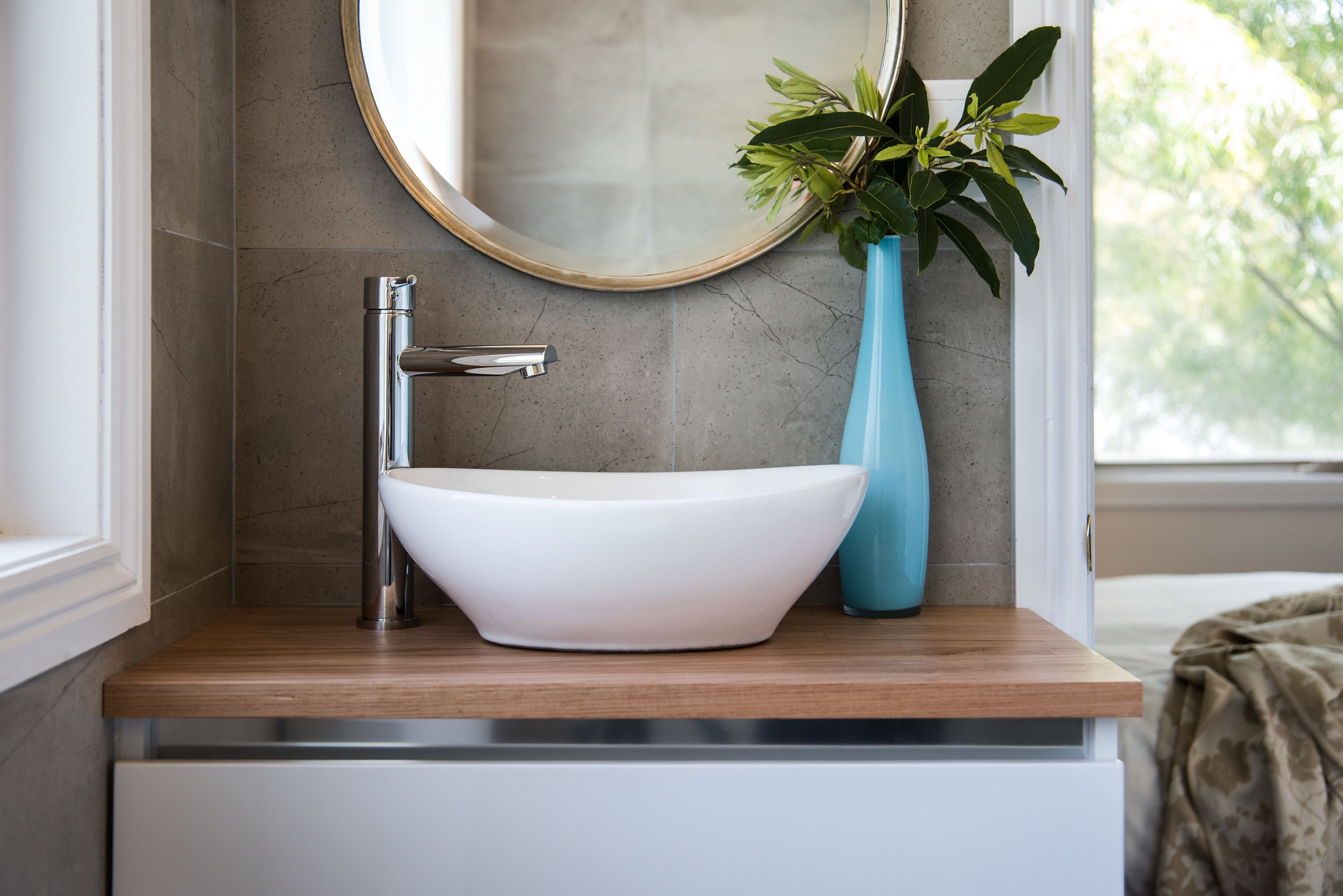 Un baño con un lavabo blanco tipo recipiente, un grifo moderno y un jarrón azul con hojas verdes.