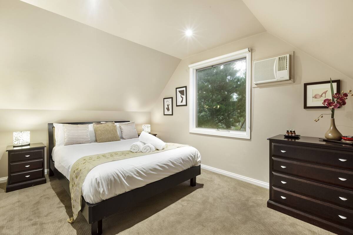 Une chambre à coucher comprenant un lit, des tables de chevet, une commode et une fenêtre donnant sur des arbres.