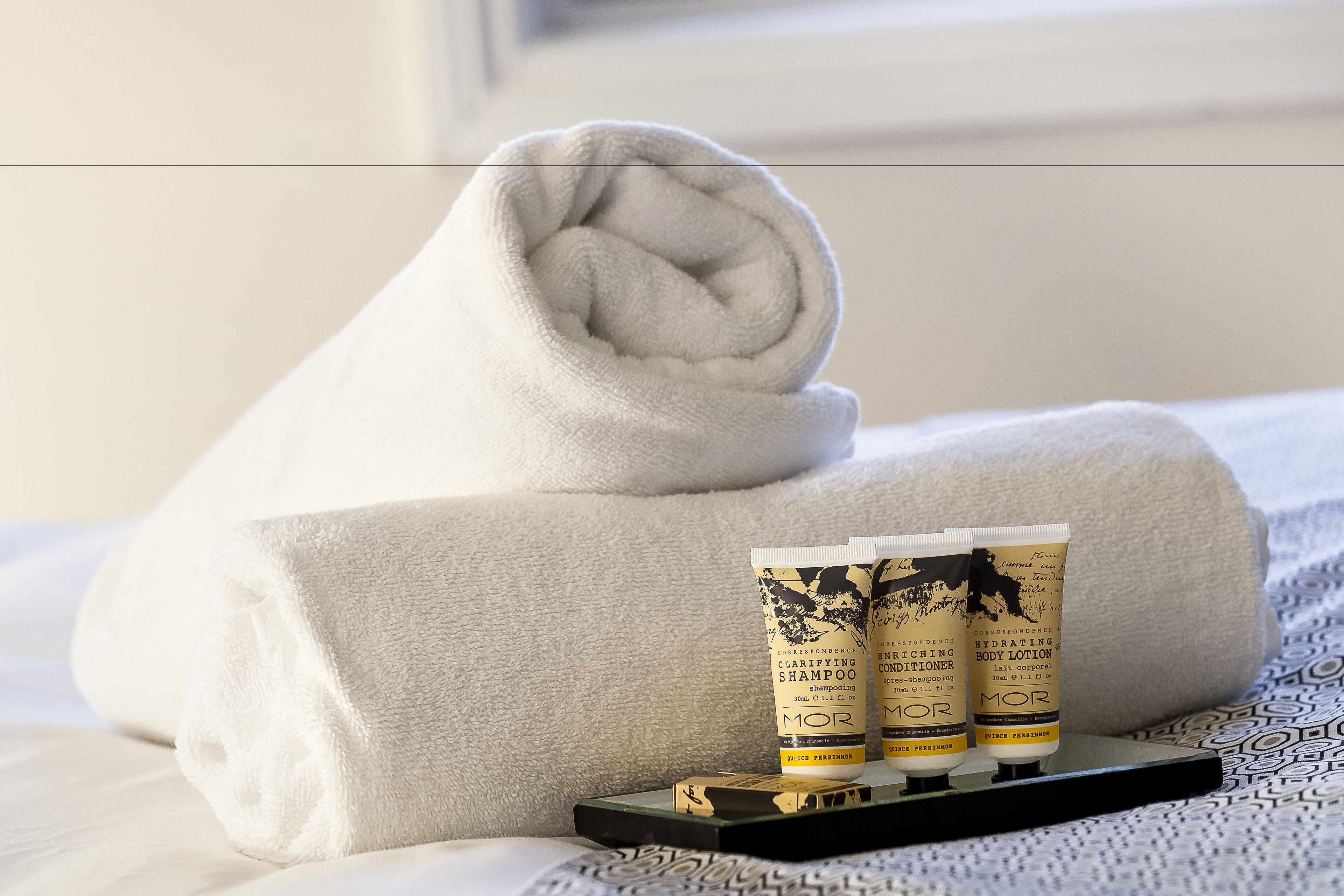 A neatly folded white towel, two bottles of Clarifying Shampoo, and a box of Hydrating Body Lotion on a bed with patterned bedding.