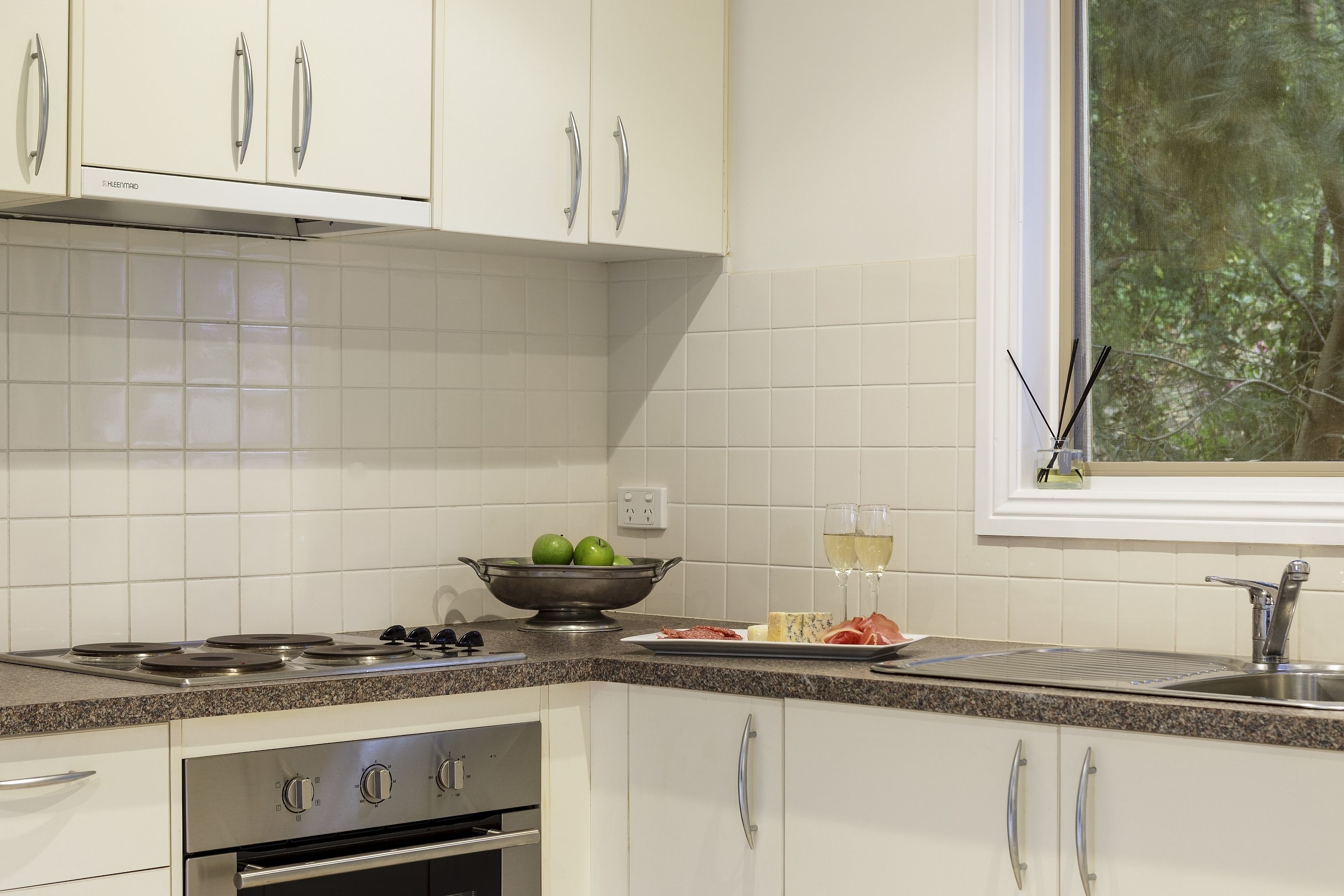 A modern kitchen with white cabinets, a stainless steel range hood, a granite countertop, and a window with a view of trees.