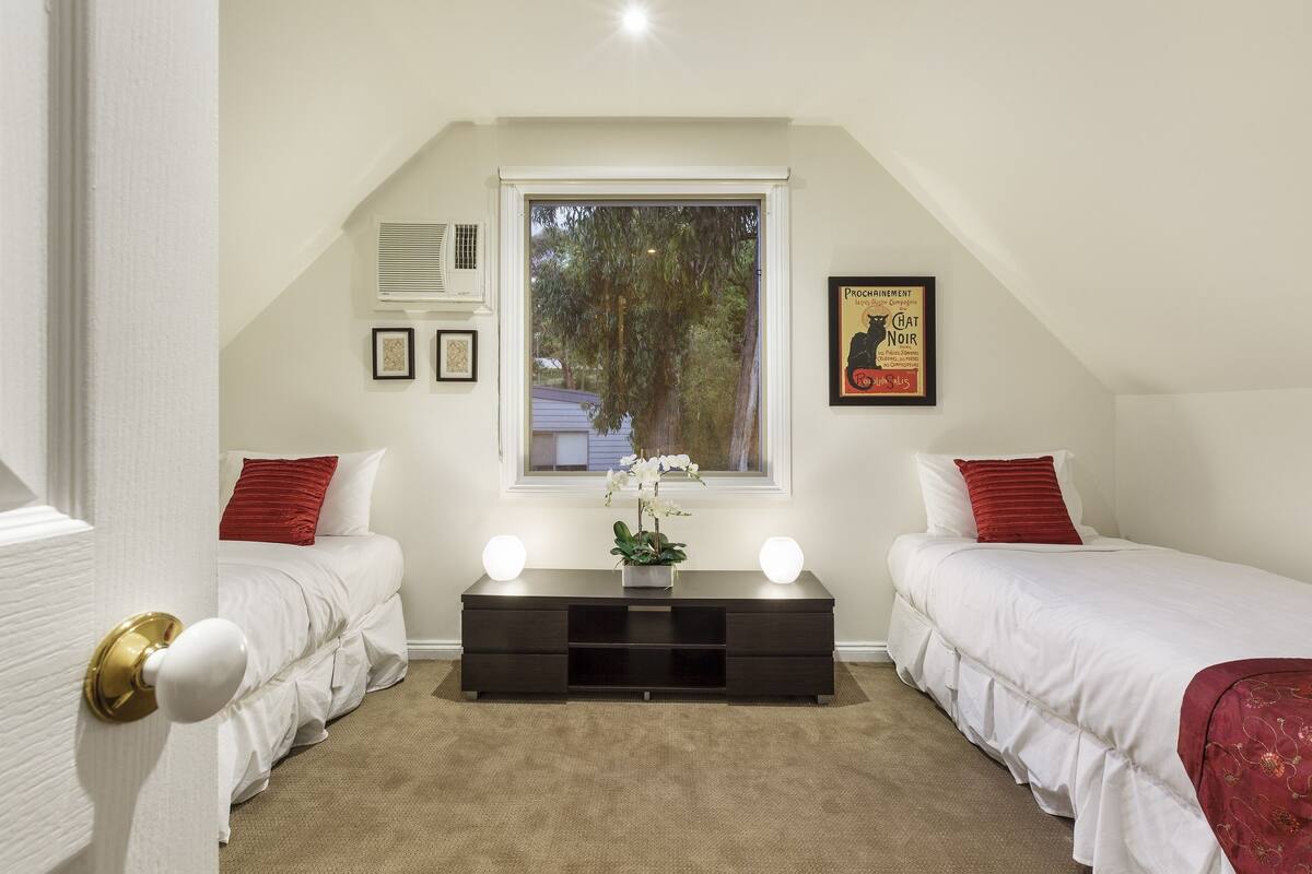 Une chambre avec deux lits simples, une commode en bois foncé, une petite table sur laquelle est posé un vase de fleurs et une fenêtre donnant sur des arbres.