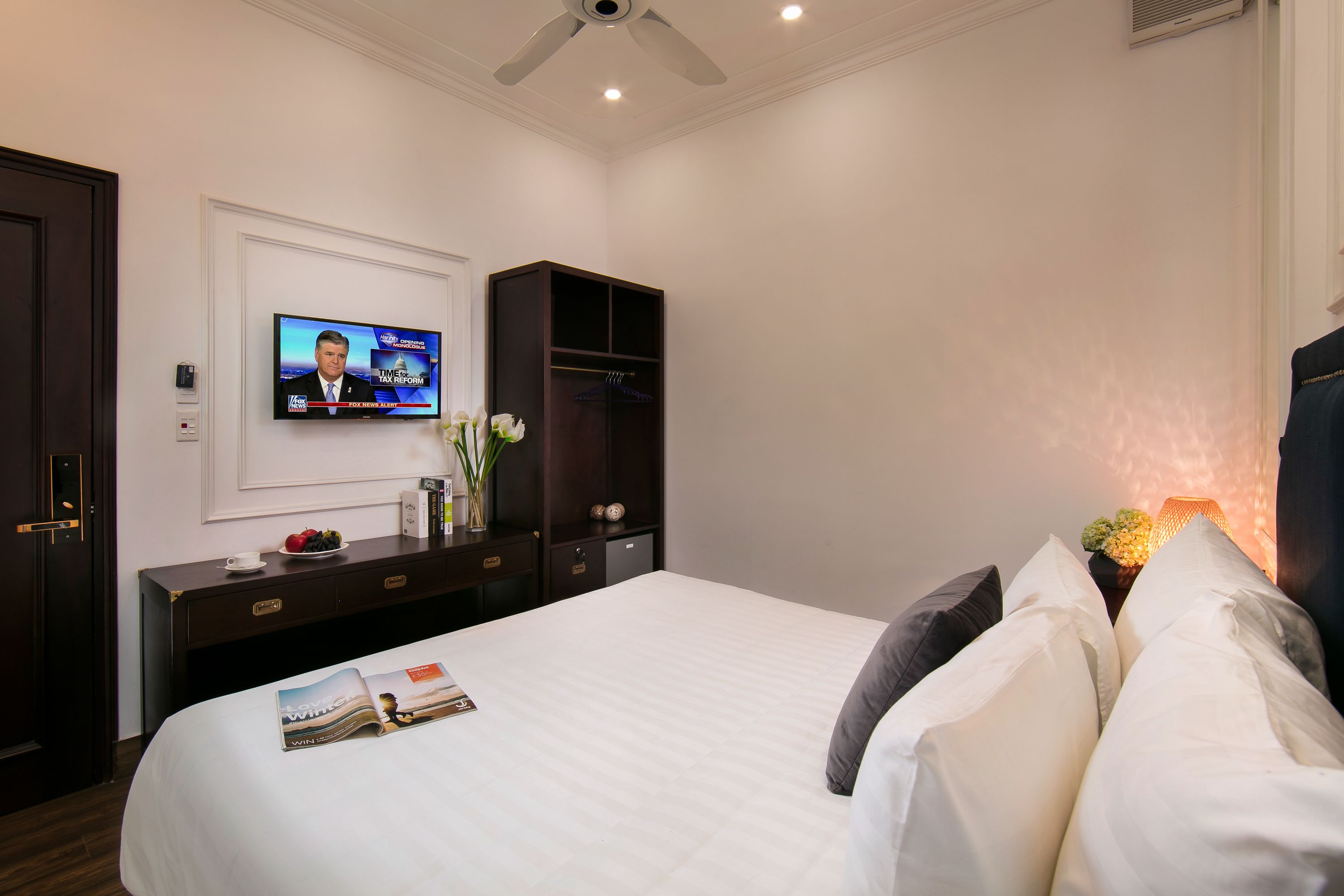 A hotel room with a bed, a TV mounted on the wall, a dark wooden dresser, and a nightstand with a lamp.