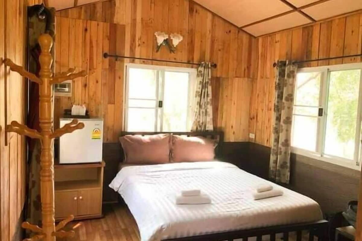 A wooden cabin bedroom with a bed, a small dresser, a coat rack, and a window with curtains.