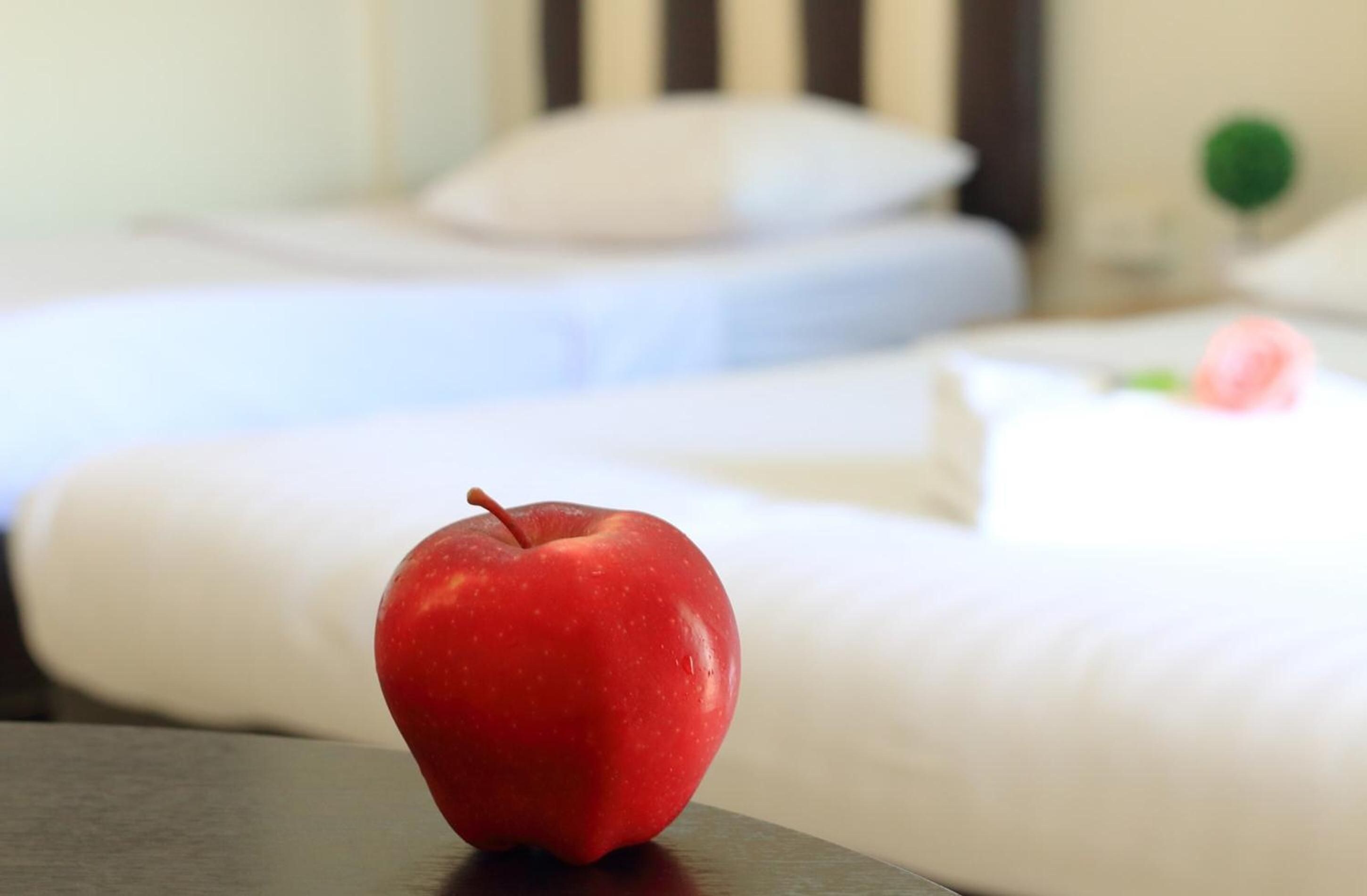 A red apple on a table in a hotel room with white bedding and a green object in the background.