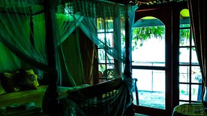 A room with a bed under a canopy, a window with a view of greenery, and a small table with a chair. - Mahi Villa (Bentota)