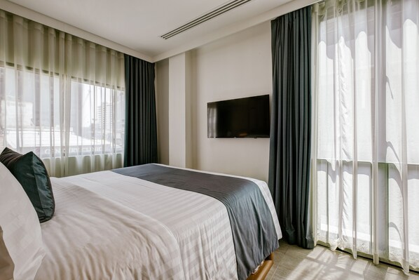 A hotel room with a large bed, a flat-screen TV mounted on the wall, and a window with sheer curtains.