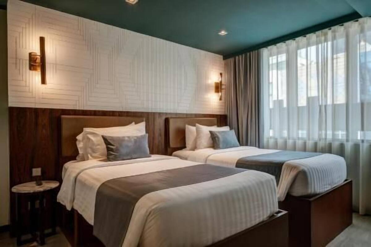 A hotel room with two beds, a round table, and a view of the outdoors through sheer curtains.