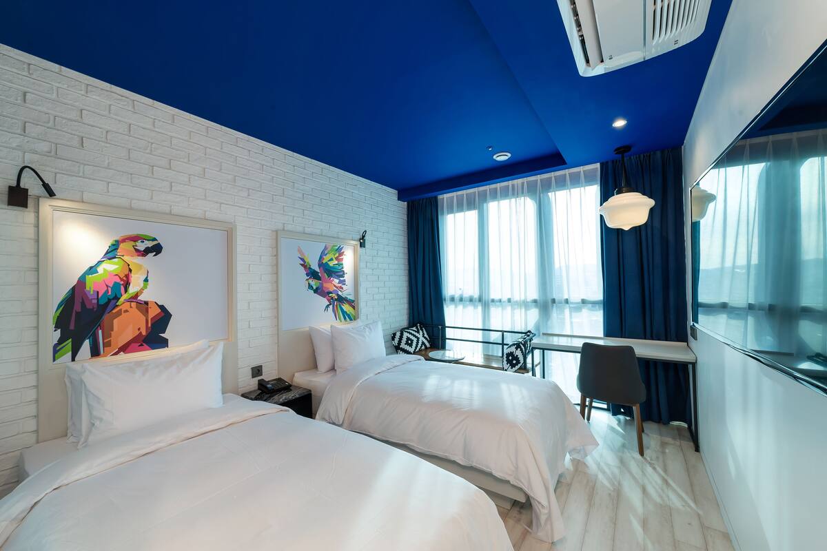 A modern hotel room with two beds, a white brick wall, a blue ceiling, and large windows with blue curtains.