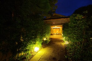 A traditional Japanese building at night with a pathway leading to the entrance, surrounded by bamboo plants and illuminated by warm lighting.