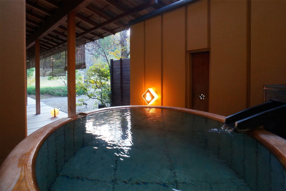 A hot tub with a wooden frame and a water feature, surrounded by a wooden deck and a covered area with a view of a garden and trees.