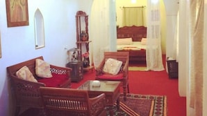Una habitación con una cama, dos sillas, una mesita y un espejo. Hay un cuadro en la pared y una ventana con cortinas.