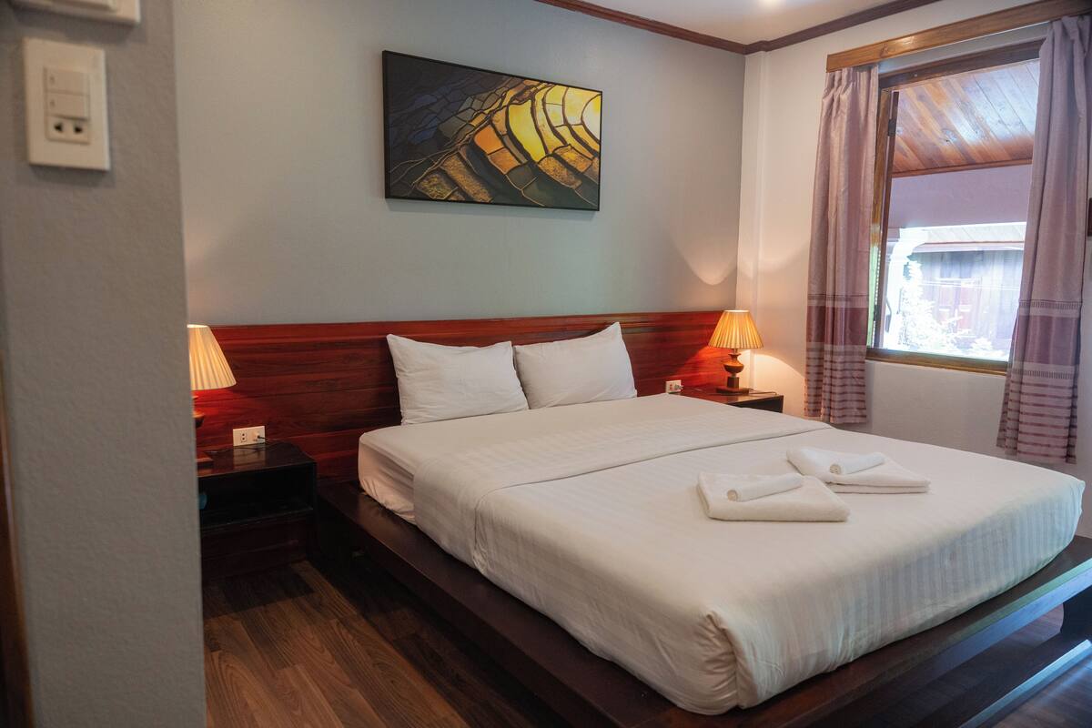 Een netjes opgemaakt bed met witte lakens en twee kussens, een houten hoofdbord, een nachtkastje met een lamp en een schilderij aan de muur.