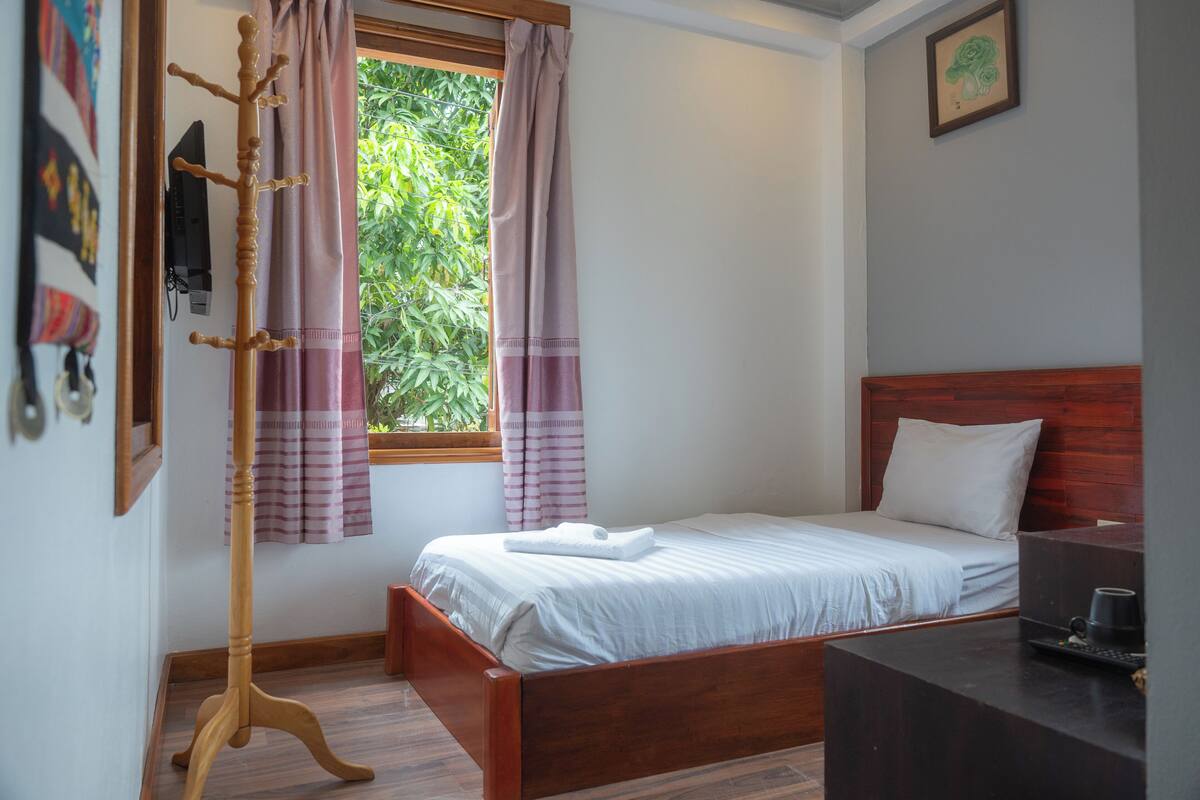 Een netjes opgemaakte slaapkamer met een houten bed, nachtkastje, televisie en een raam met gordijnen.