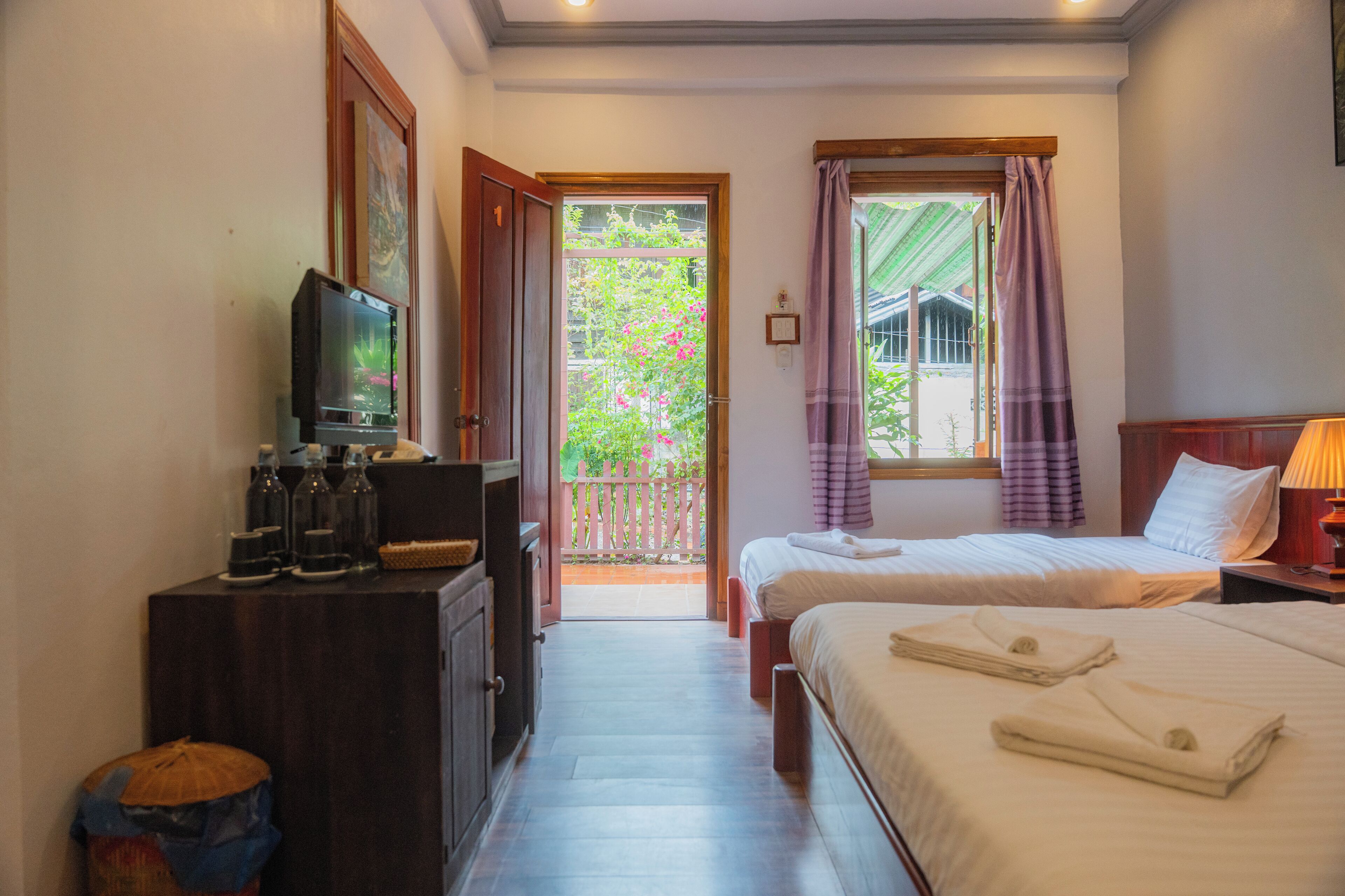 A hotel room with two beds, a TV, and a balcony with a view of greenery.