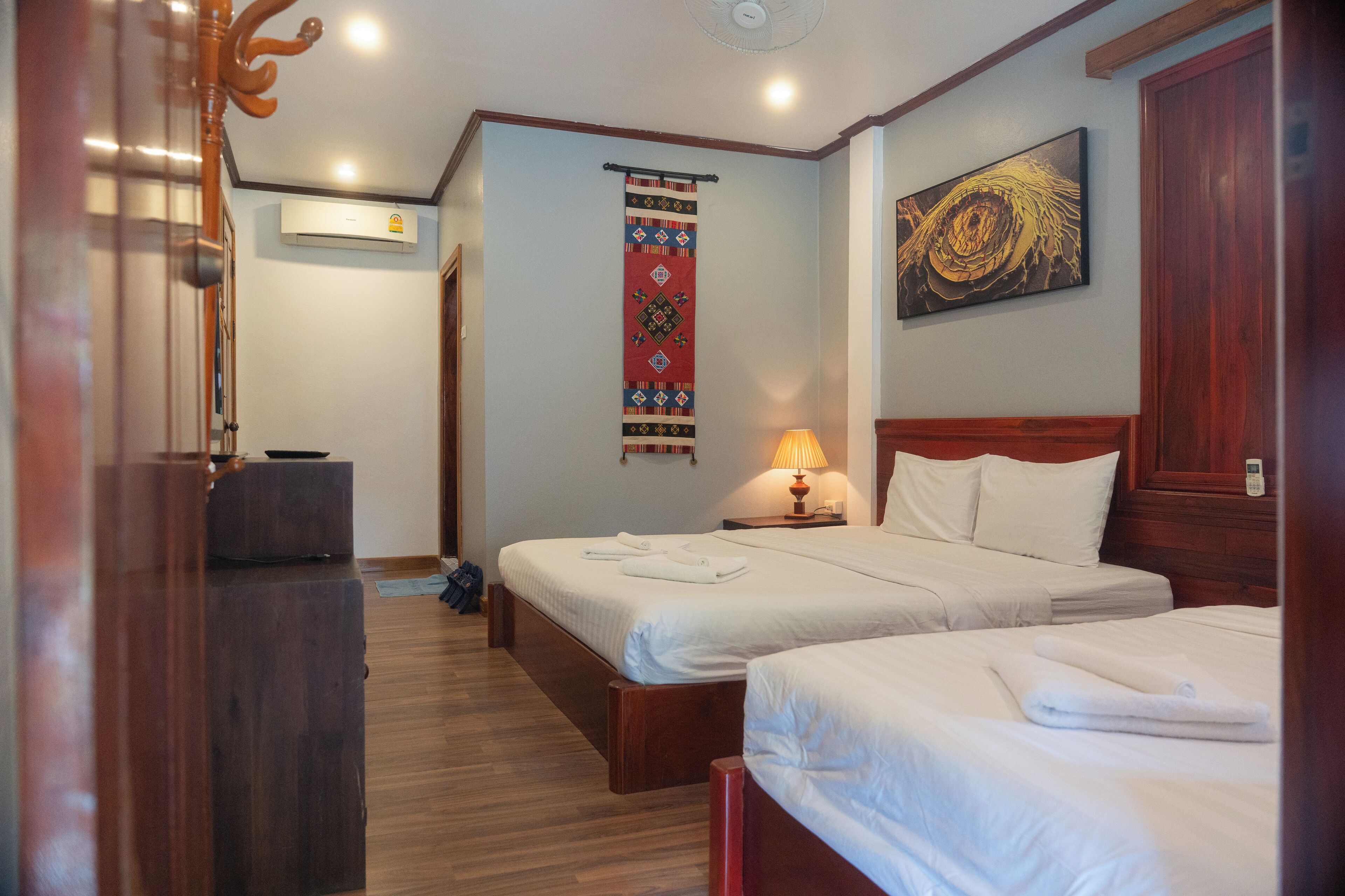 A hotel room with two beds, a wooden headboard, a wall-mounted air conditioner, a decorative hanging, and a painting.