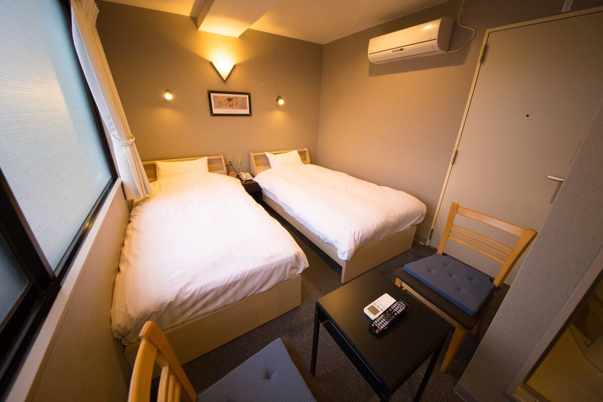 A small hotel room with two single beds, a small table with a remote control, a chair, and an air conditioning unit on the wall.