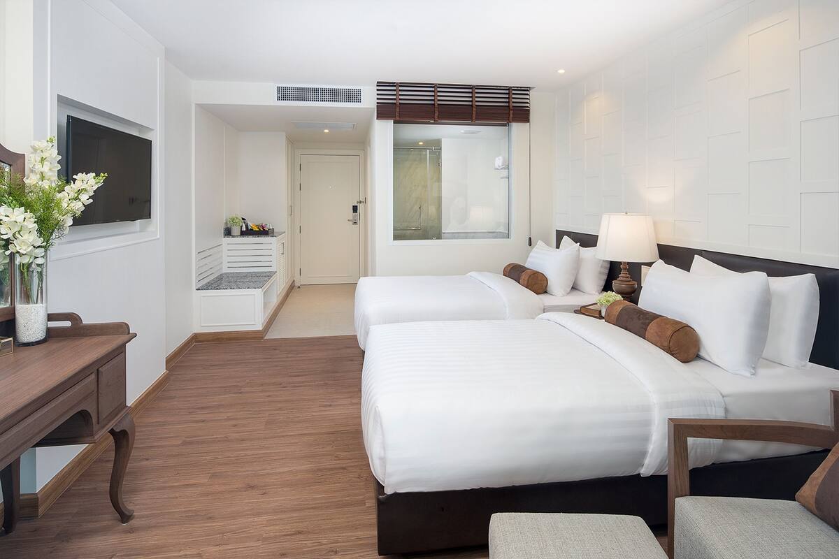 A hotel room with two beds, a desk, a chair, a TV, and a window with a view of the interior.