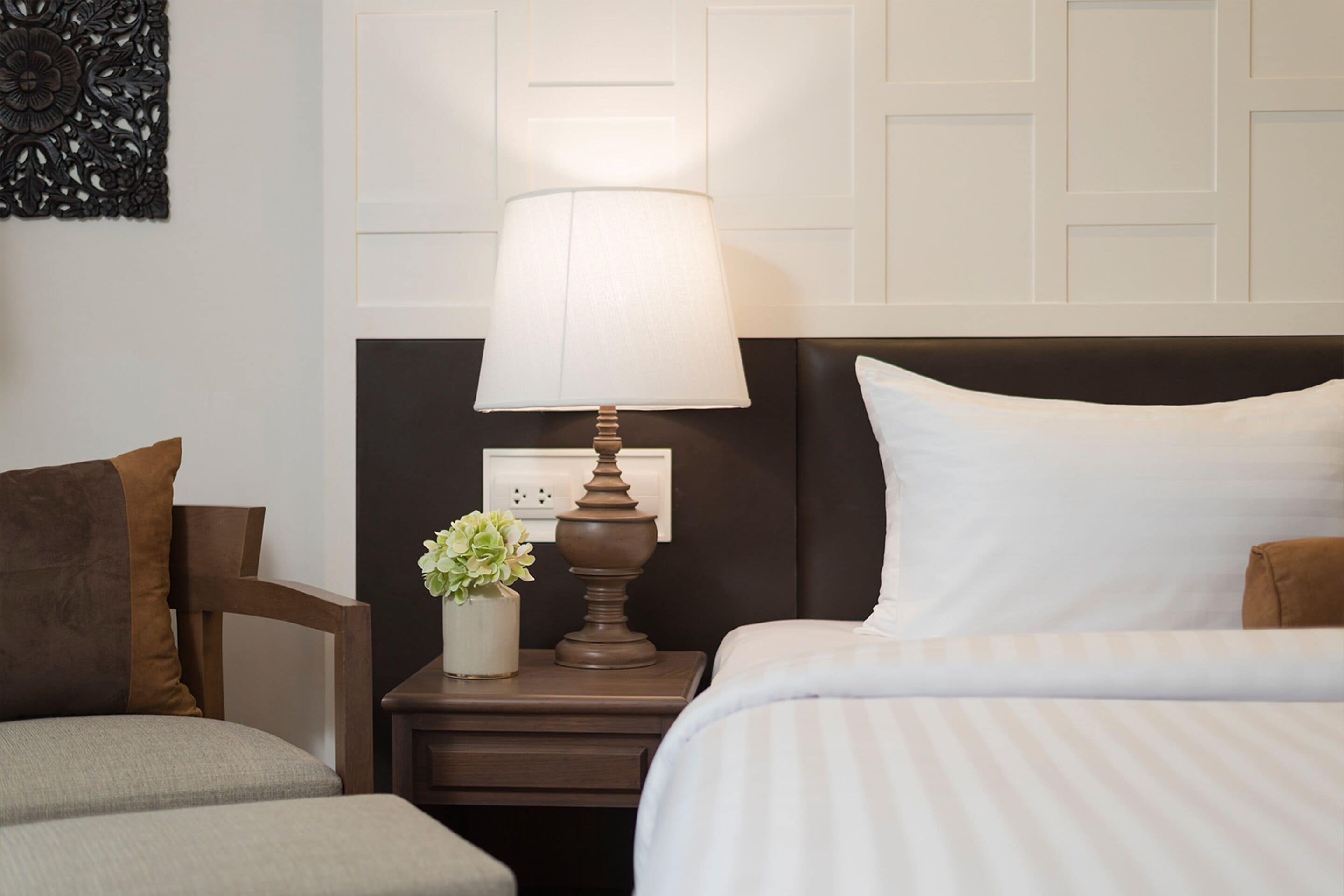 Habitación de hotel con una cama, una mesita de noche con lámpara, un jarrón con flores y un adorno en la pared.