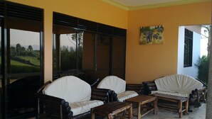 A room with two wooden sofas and a small table, large windows with blinds, and a painting on the wall. - The Crested Crane Bwindi (Kyesero)