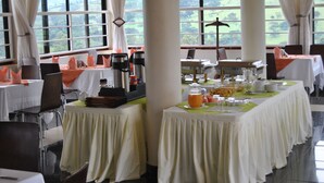 A dining area with tables set for breakfast, featuring orange napkins, a buffet with various food items, and a view of the outdoors through large windows. - The Crested Crane Bwindi (Kyesero)