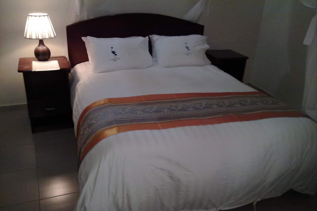 A neatly made bed with a white duvet, two pillows, and a decorative blanket. There is a bedside table with a lamp and a small drawer.