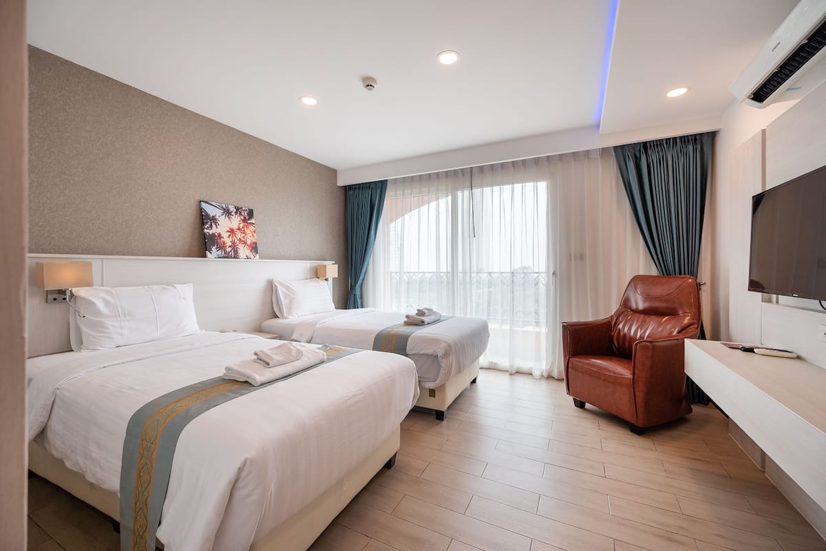 A hotel room with two beds, a TV, a leather chair, and a large window with curtains.