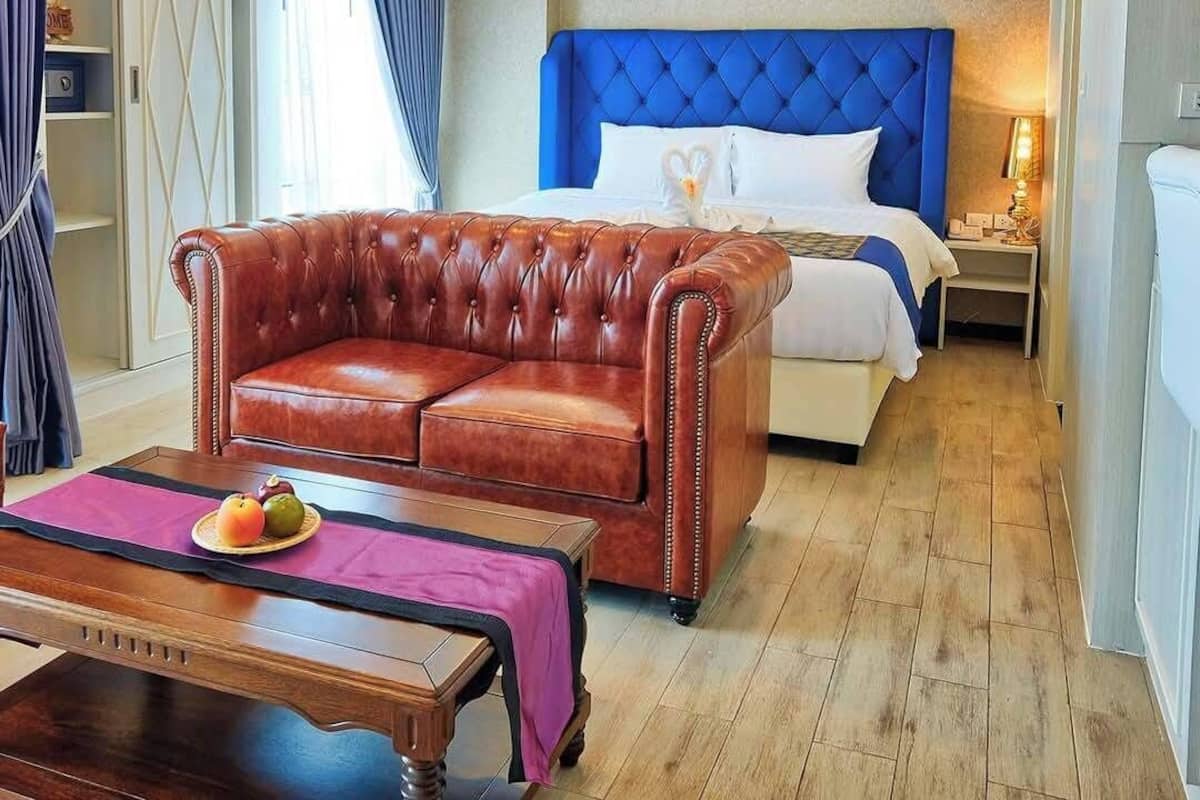A hotel room with a brown leather sofa, a wooden coffee table with fruits, a bed with a blue headboard, and a nightstand with a lamp.