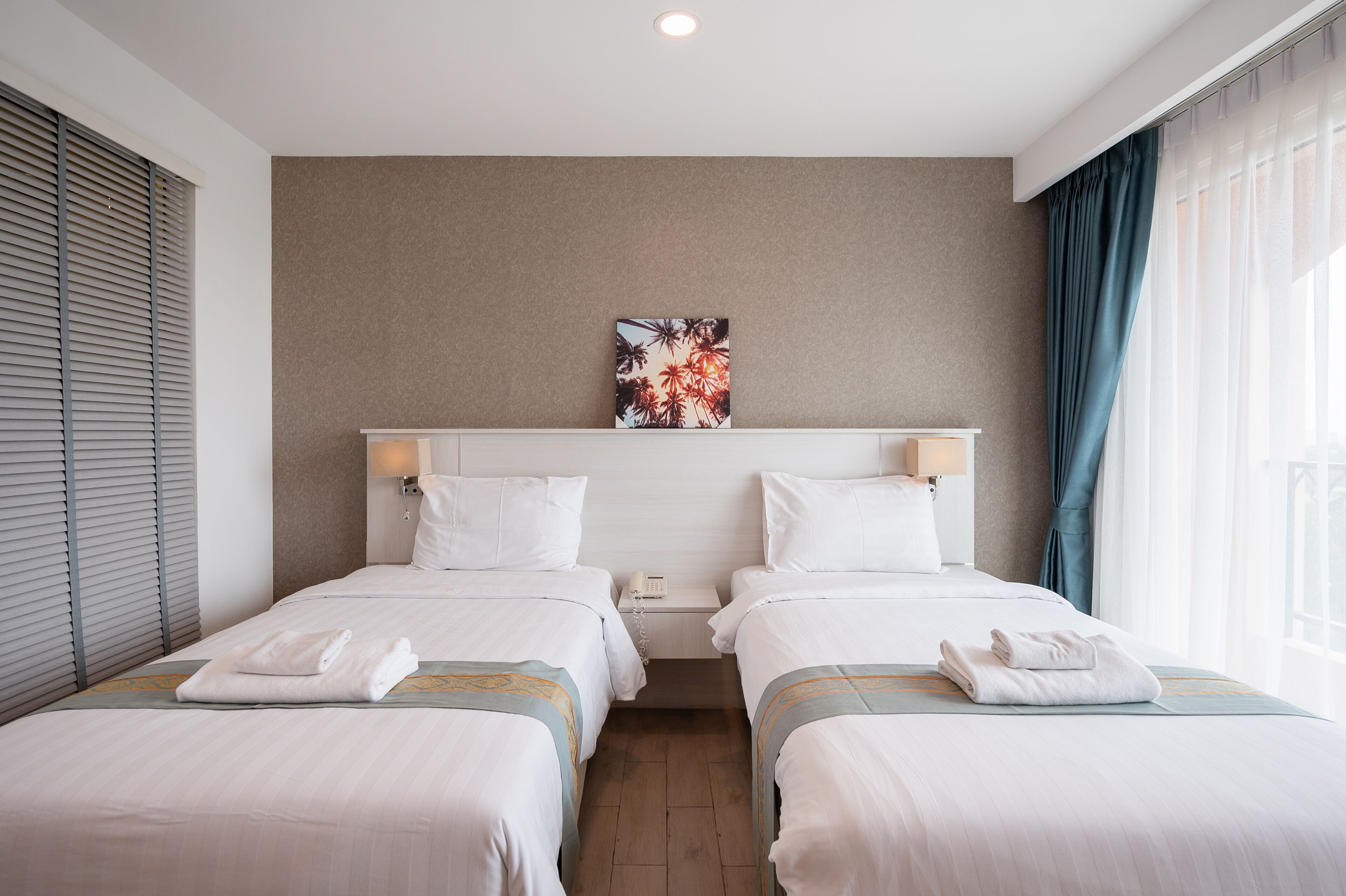 A hotel room with two beds, a headboard with a floral artwork, a bedside table with a lamp, and a window with curtains.