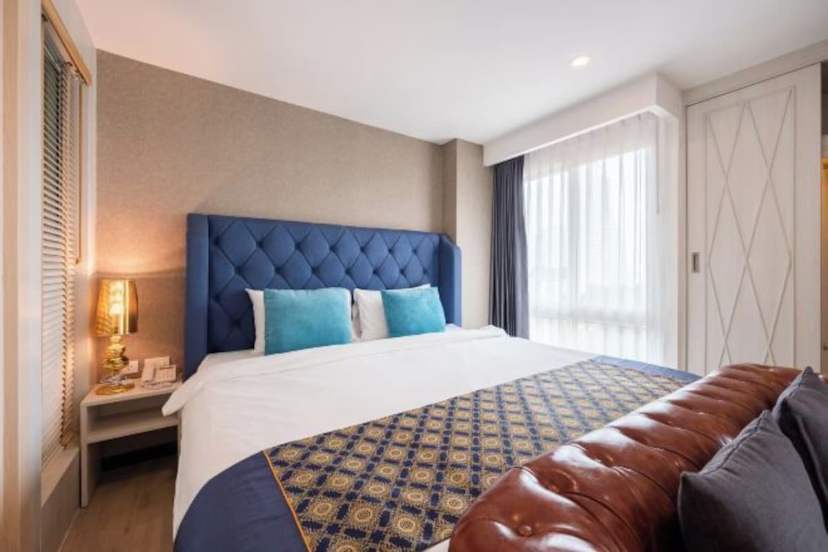 A hotel room with a large bed, a blue headboard, a patterned bedspread, a leather bench, and a nightstand with a lamp.
