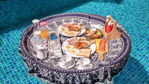 Un tavolo rotondo con tovaglia a motivi blu e bianchi, apparecchiato per la colazione con pane tostato, uova, fagioli e una ciotola di frutta, posto accanto a una piscina con piastrelle di mosaico blu.