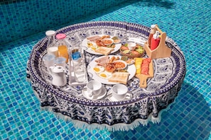 A round table with a blue and white patterned tablecloth, set with breakfast items including toast, eggs, beans, and a fruit bowl, placed by a pool with blue mosaic tiles.