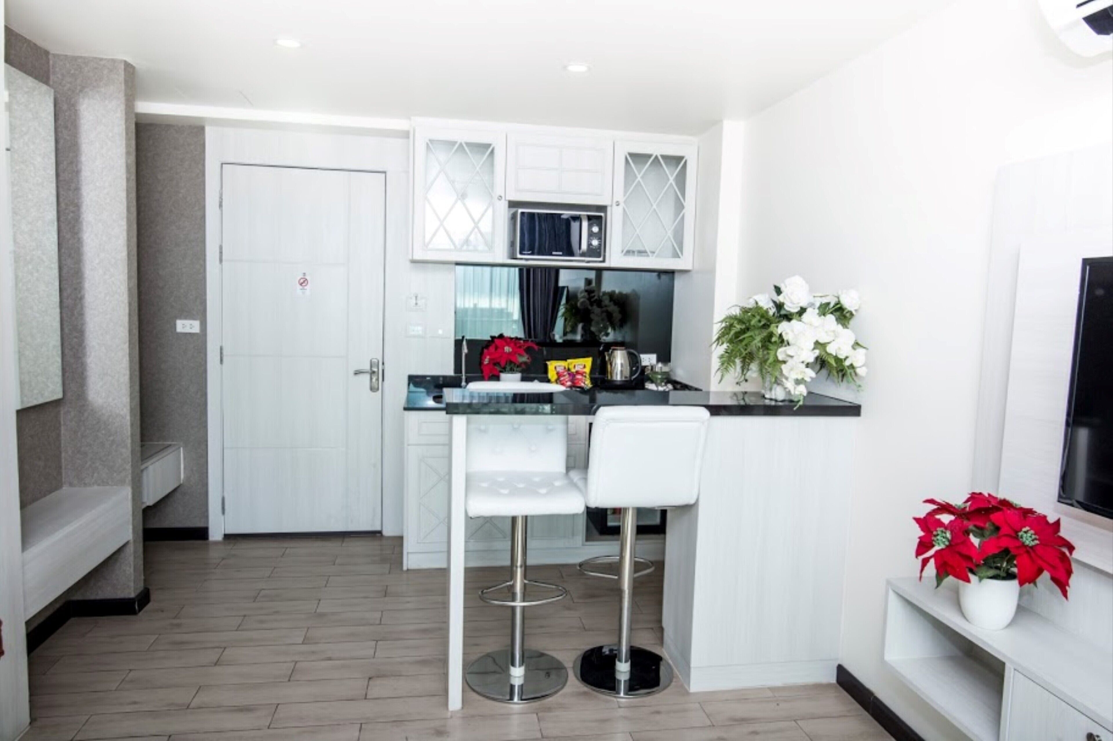 A modern kitchen with white cabinetry, a black countertop, and two white bar stools. There is a microwave above the counter, a door with a no-smoking sign, and a potted plant with red flowers.