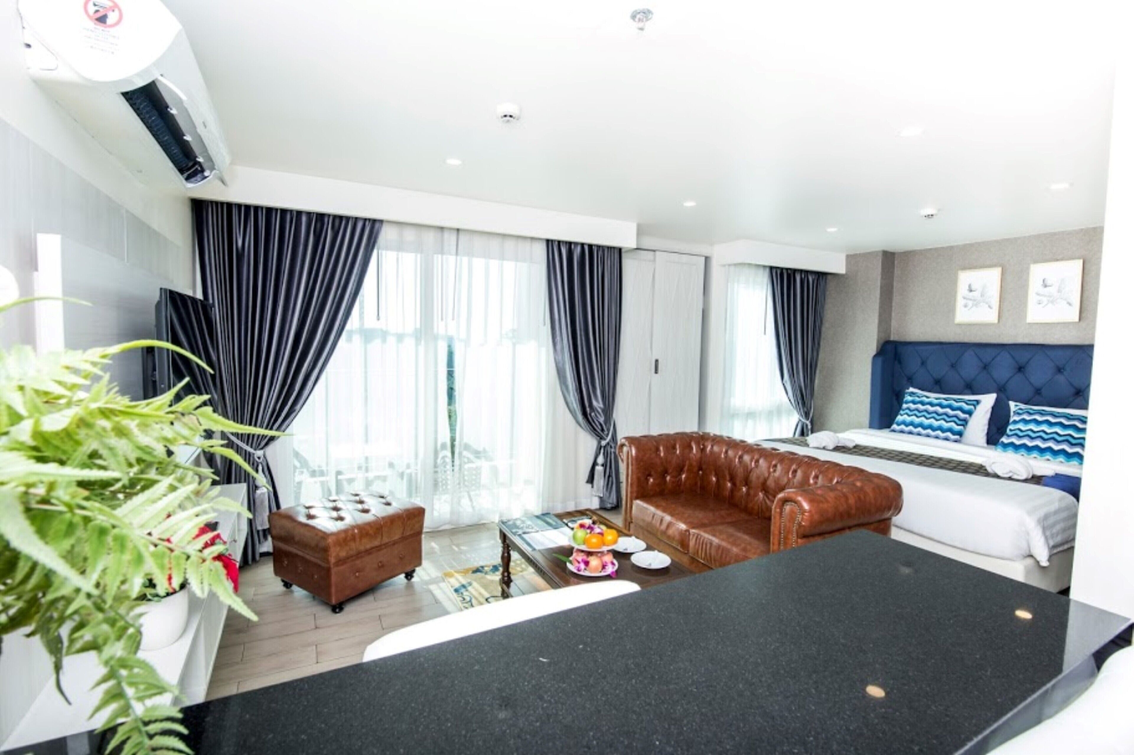 Una habitación de hotel moderna con un sofá de cuero, una cama con ropa de cama azul y blanca, una mesa de comedor y un gran ventanal con cortinas.