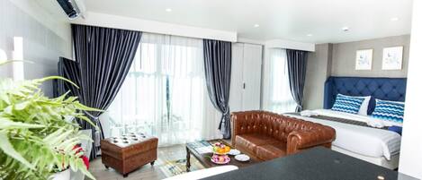 Una habitación de hotel moderna con un sofá de cuero, una cama con ropa de cama azul y blanca, una mesa de comedor y un gran ventanal con cortinas.