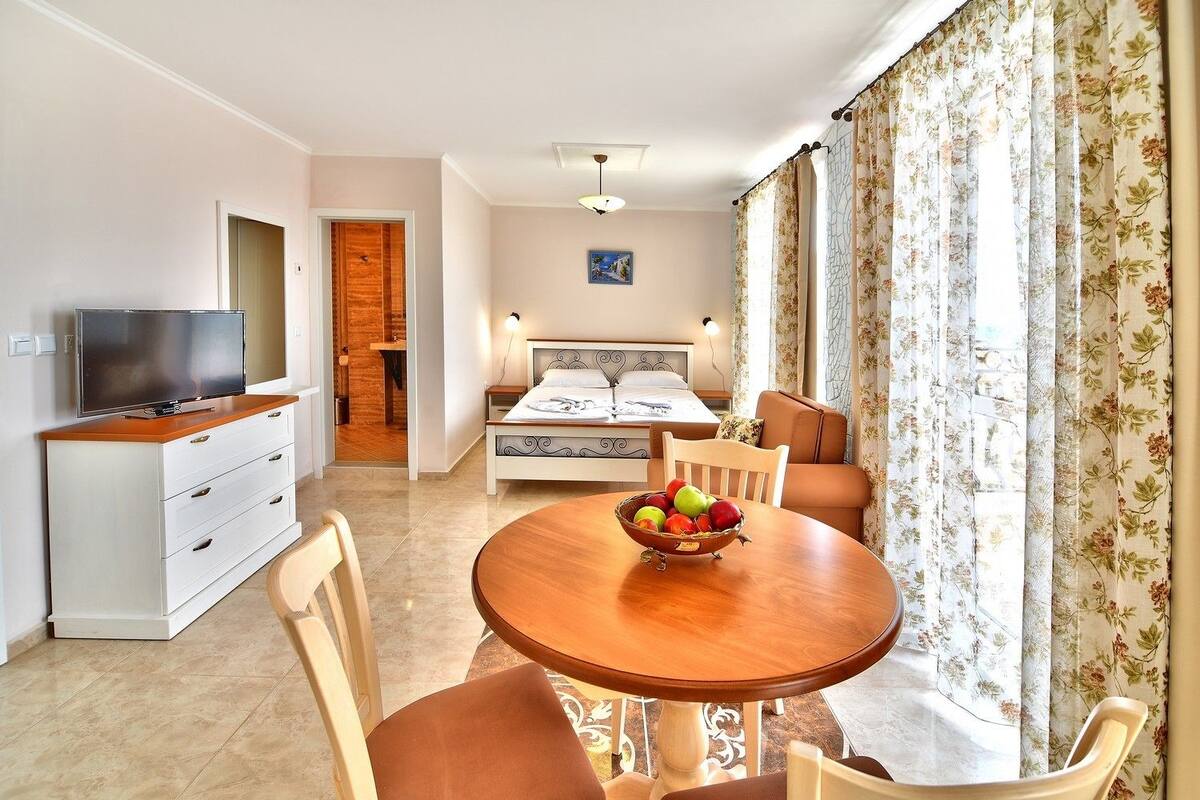 Une chambre moderne avec une table à manger en bois, un téléviseur posé sur un meuble, un lit avec une tête de lit et une fenêtre agrémentée de rideaux à motifs floraux.