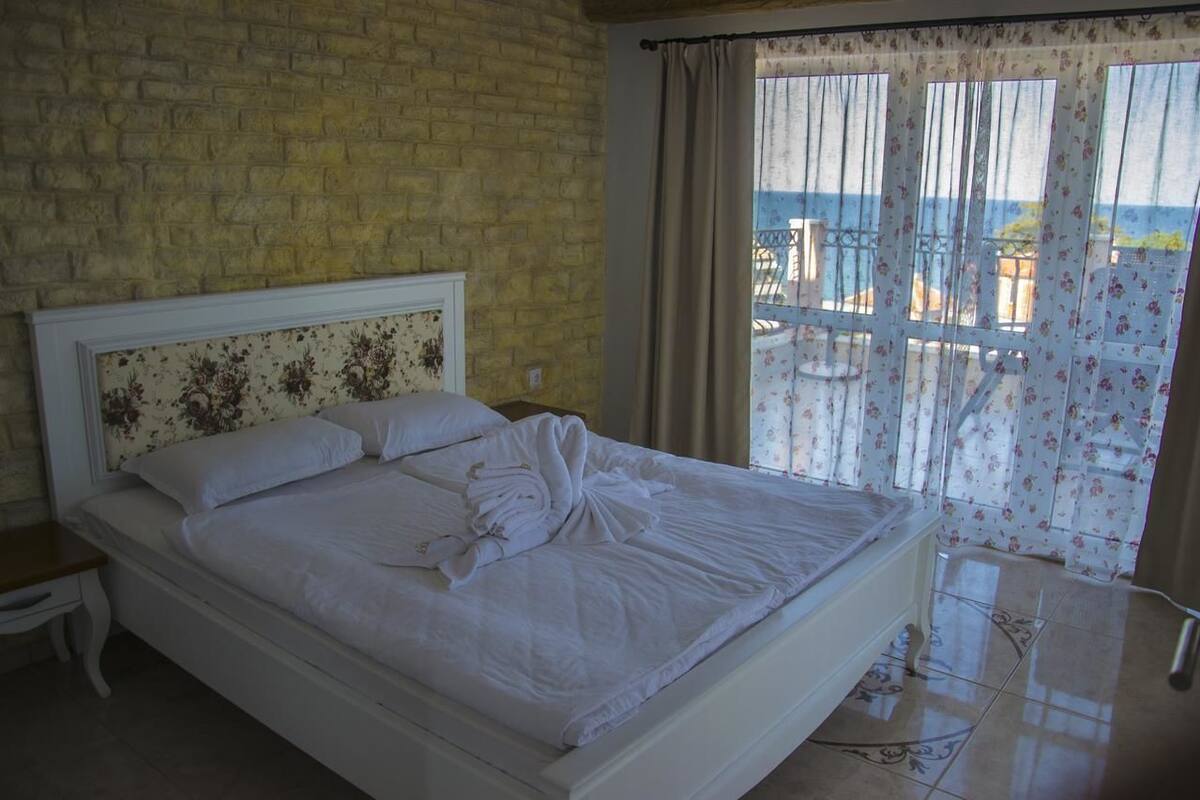 Un lit avec du linge de lit blanc et une tête de lit à motifs floraux, une table de chevet et une vue sur la mer à travers les rideaux.