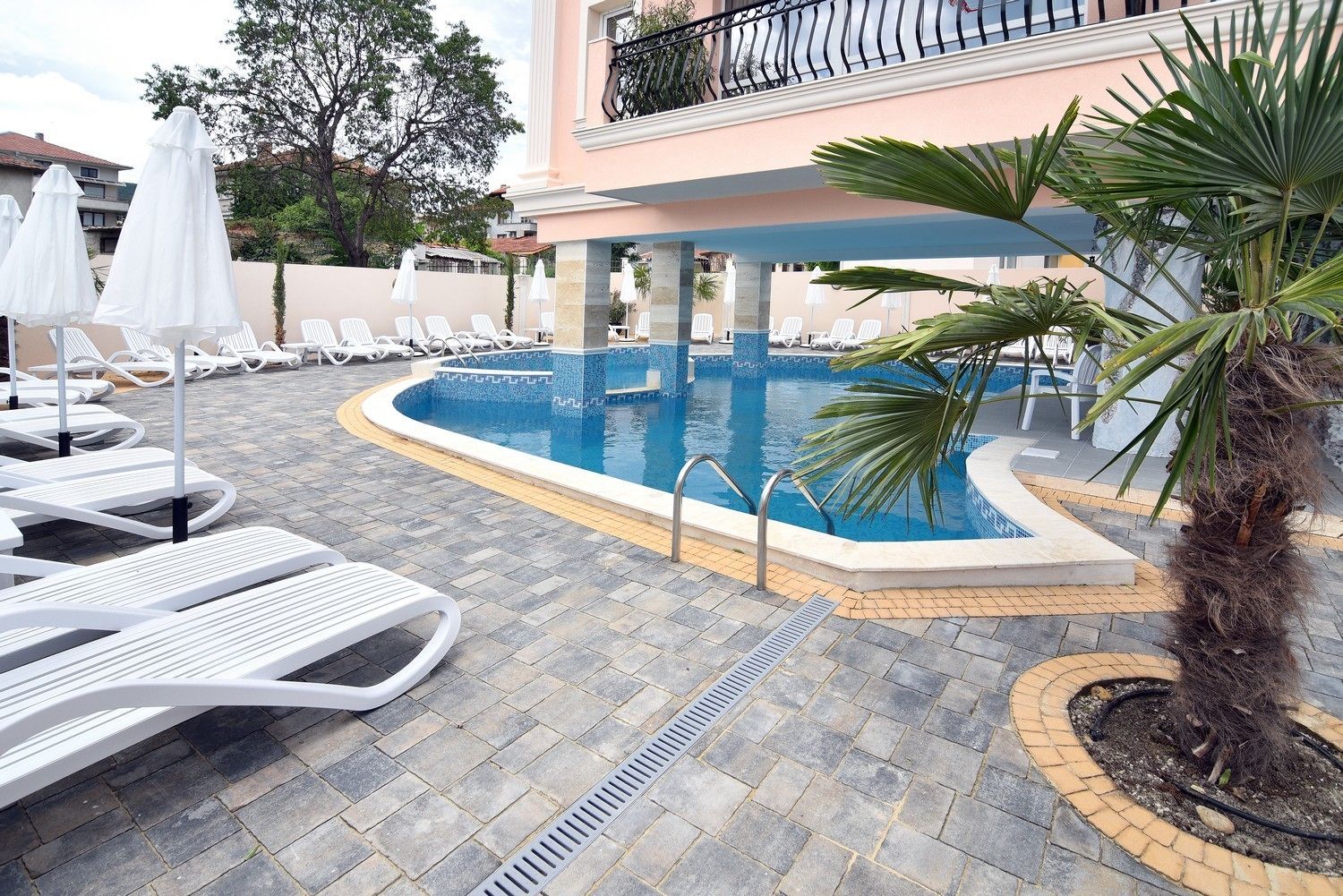 Zona de piscina con sillones de descanso y sombrillas, una palmera y un edificio al fondo.
