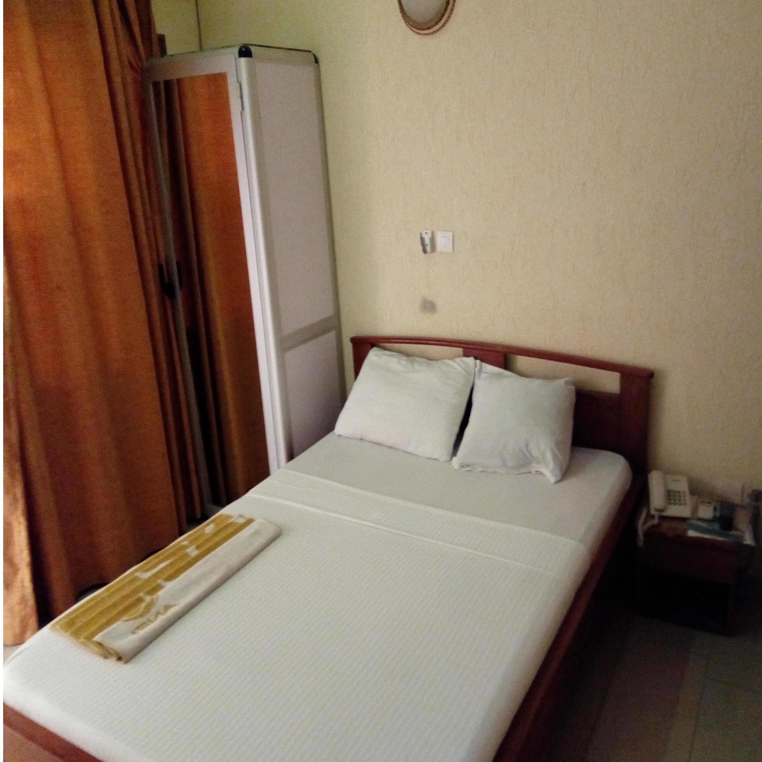 A hotel room with a bed, a bedside table with a phone, and a wall-mounted light switch.