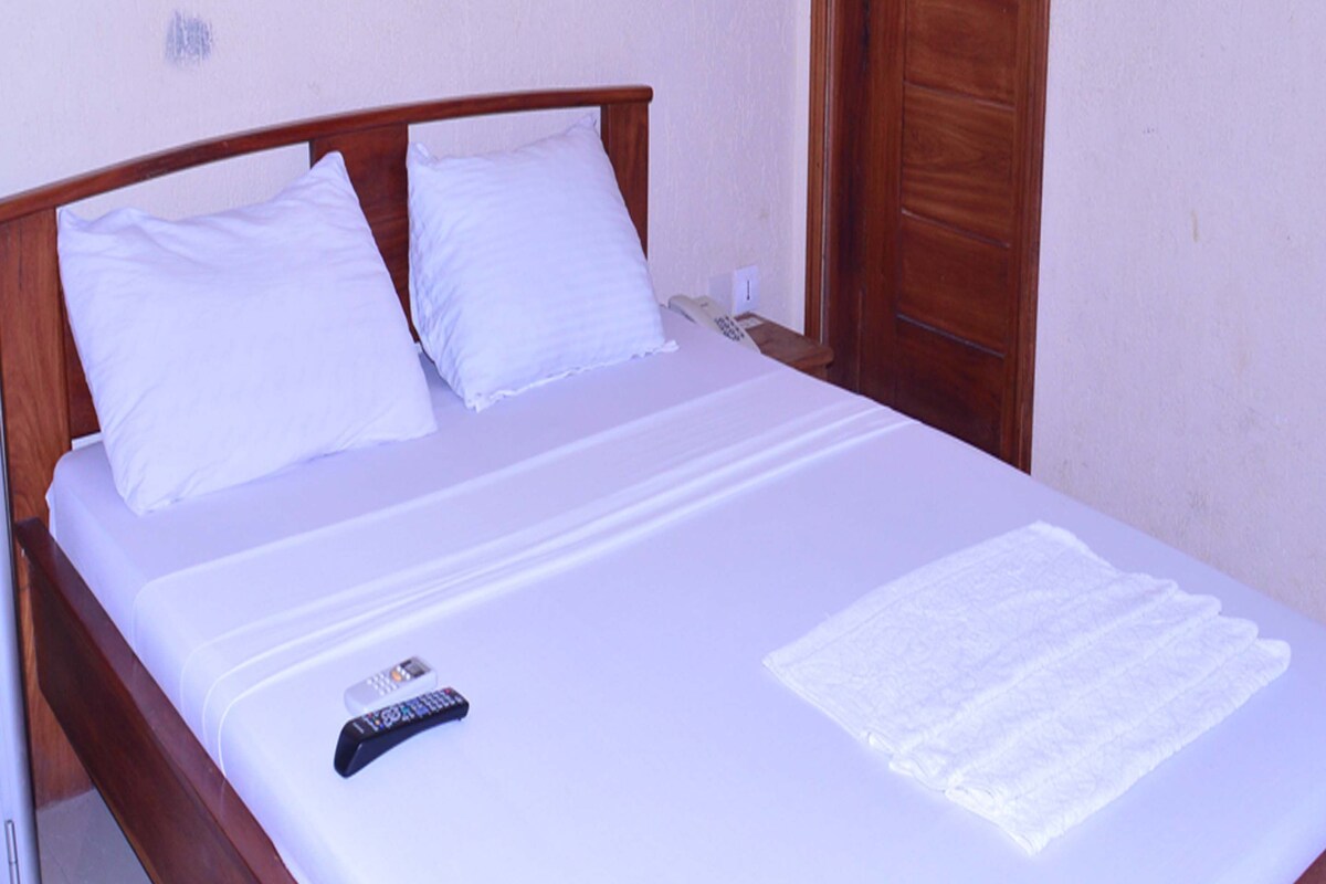 A neatly made bed with white linens, a remote control, and a folded white towel on a wooden bed frame in a room with a wooden door and light-colored walls.