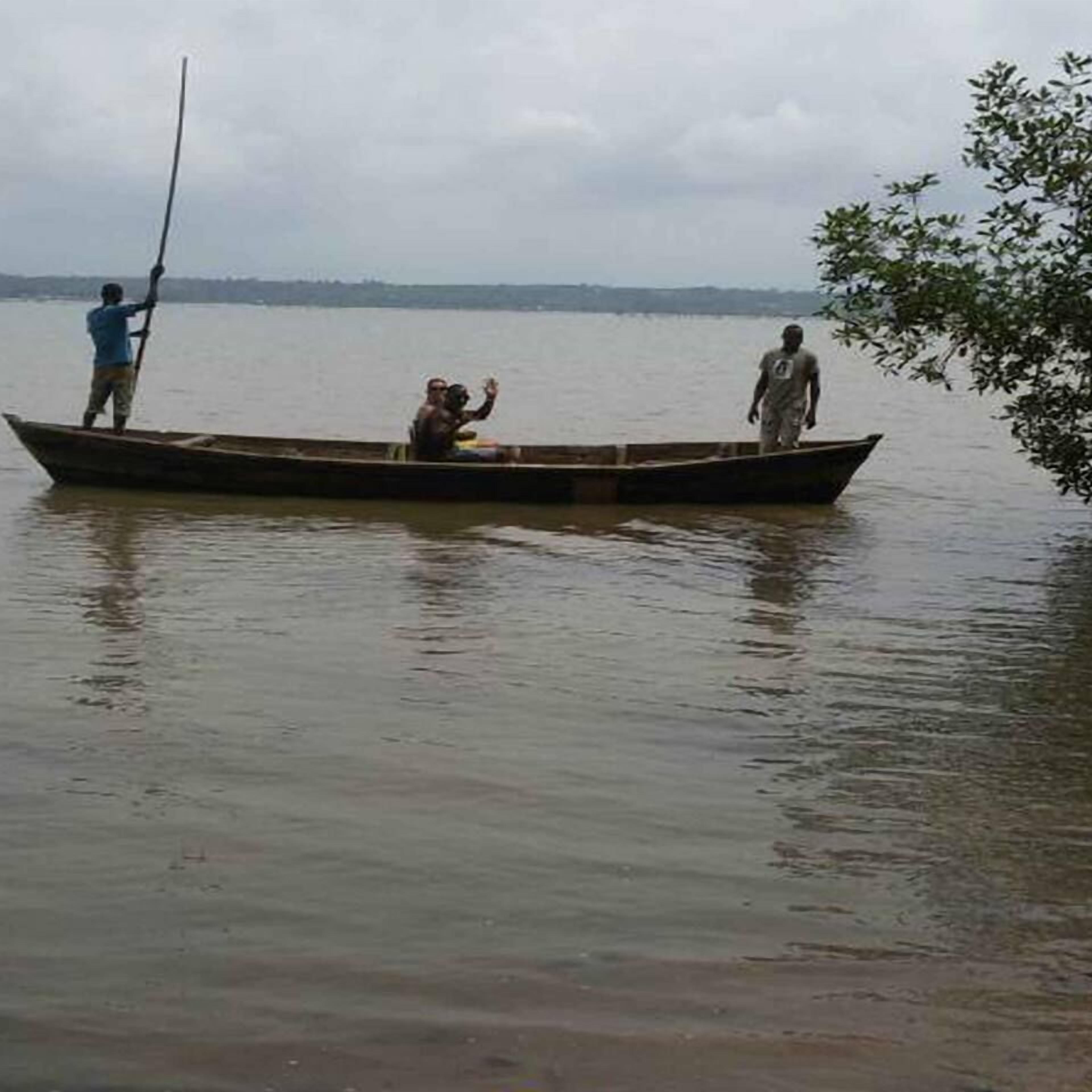Un petit bateau en bois sur une eau calme, avec trois personnes à bord : une personne se tient à l’avant, une autre à l’arrière et une troisième est assise au milieu.