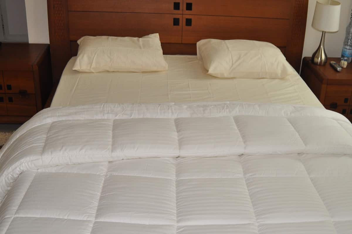 Una cama doble con edredón blanco y dos almohadas, cabecera de madera y mesita de noche con lámpara y control remoto.