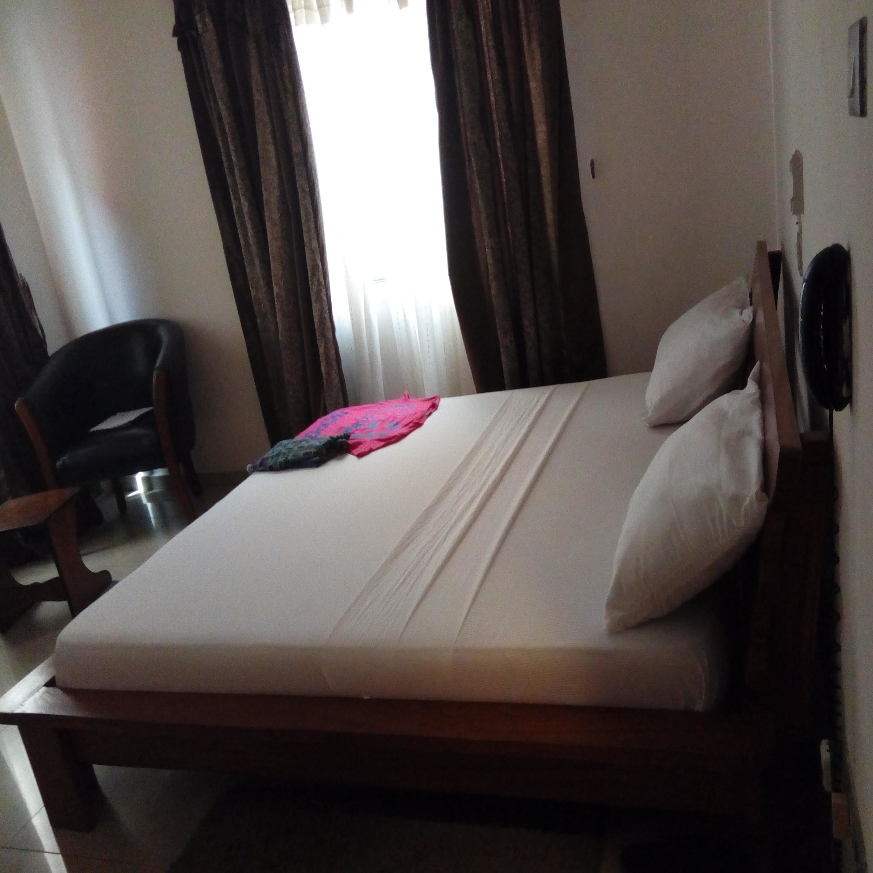 A single bed with white linens, a wooden headboard, and two pillows. There is a chair to the left and a window with curtains partially drawn.
