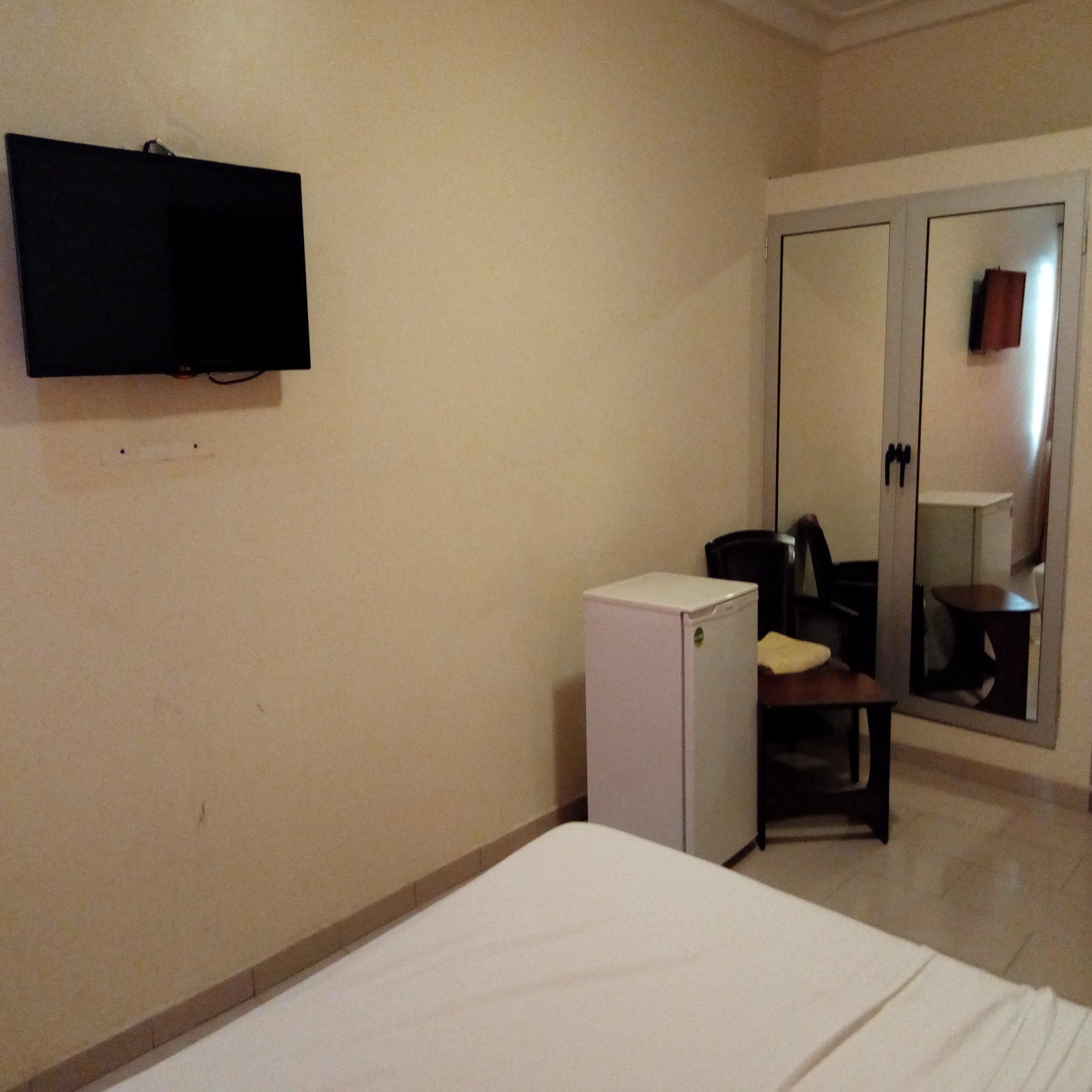 A hotel room with a bed, a small white refrigerator, a television mounted on the wall, and a mirror in the door.